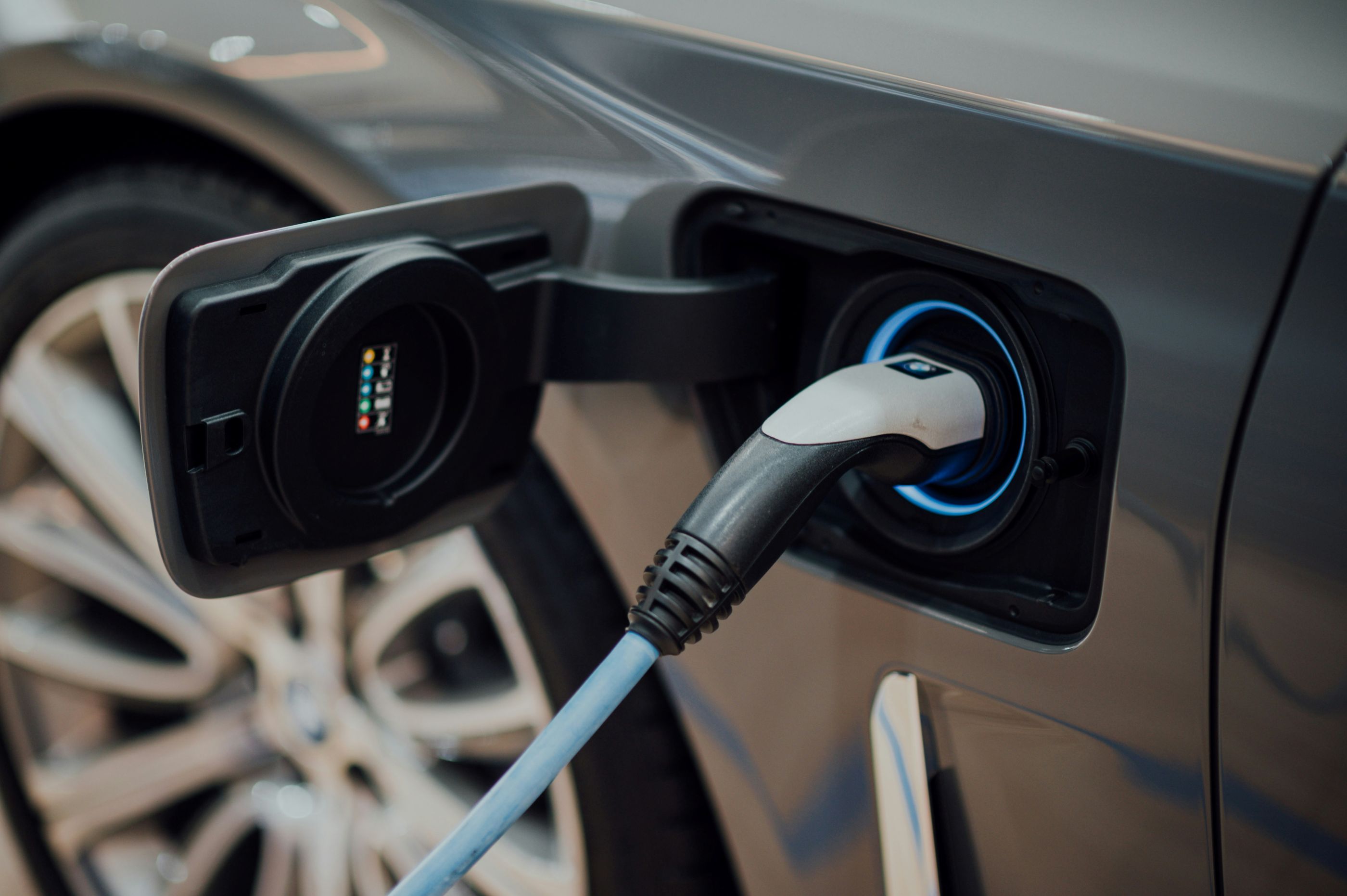 Close-up of an electric vehicle being charged, with a charging cable plugged into the port on the side of a gray car.

