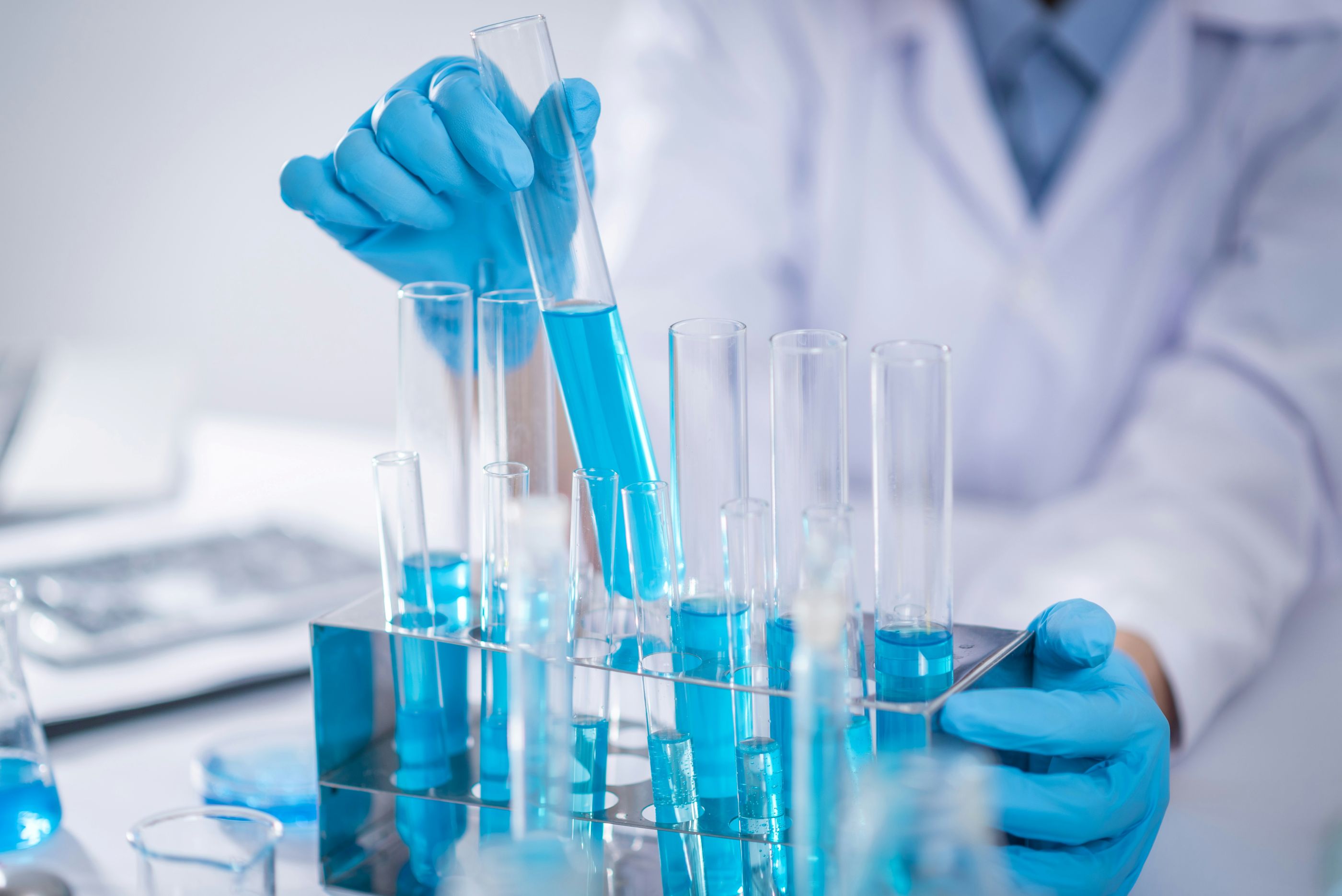 Scientist in lab coat and gloves handling test tubes filled with blue liquid in a laboratory setting, focusing on chemical analysis or pharmaceutical research.

