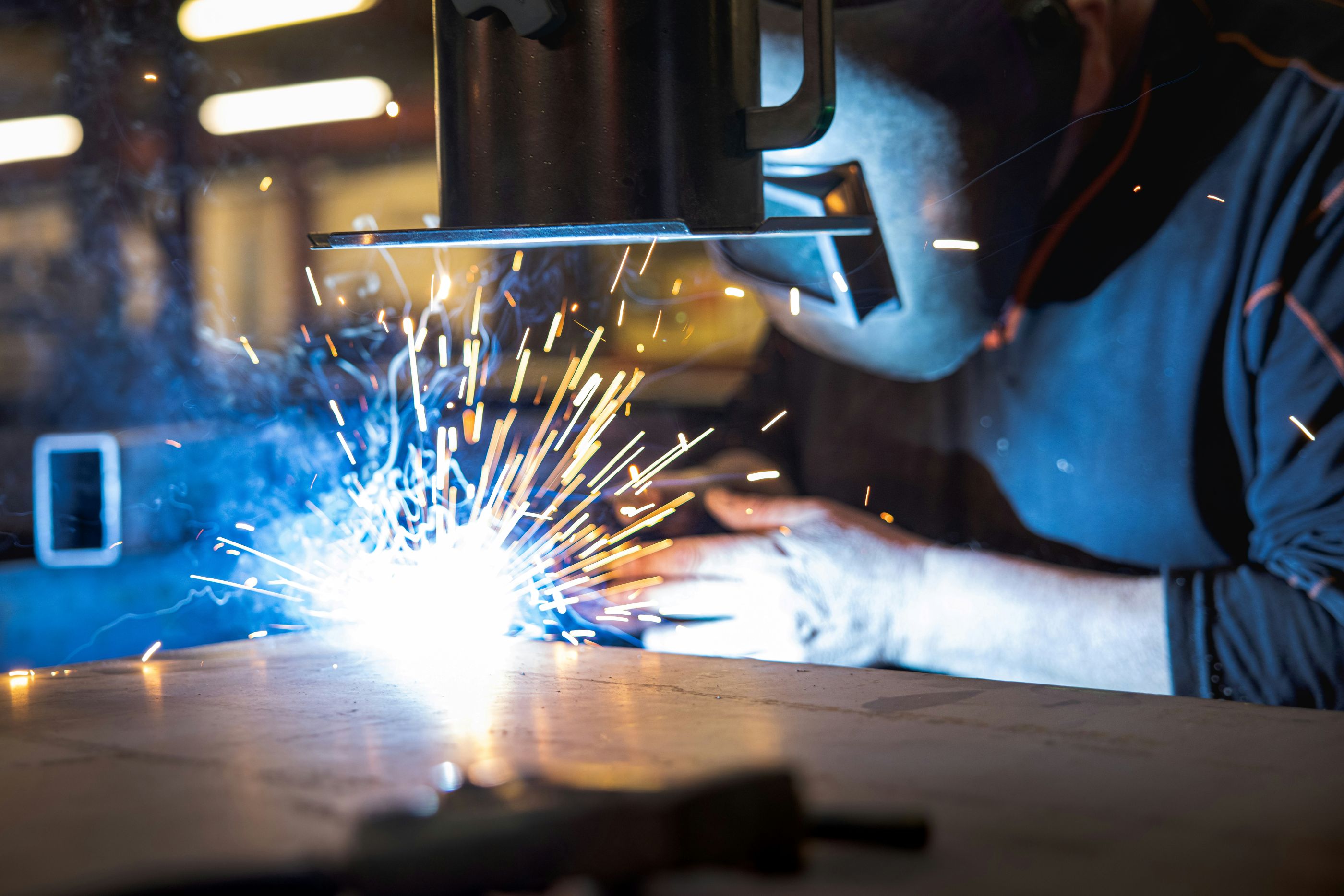 Worker wearing a protective helmet performing welding on metal, with bright sparks flying in a manufacturing or industrial workshop.

