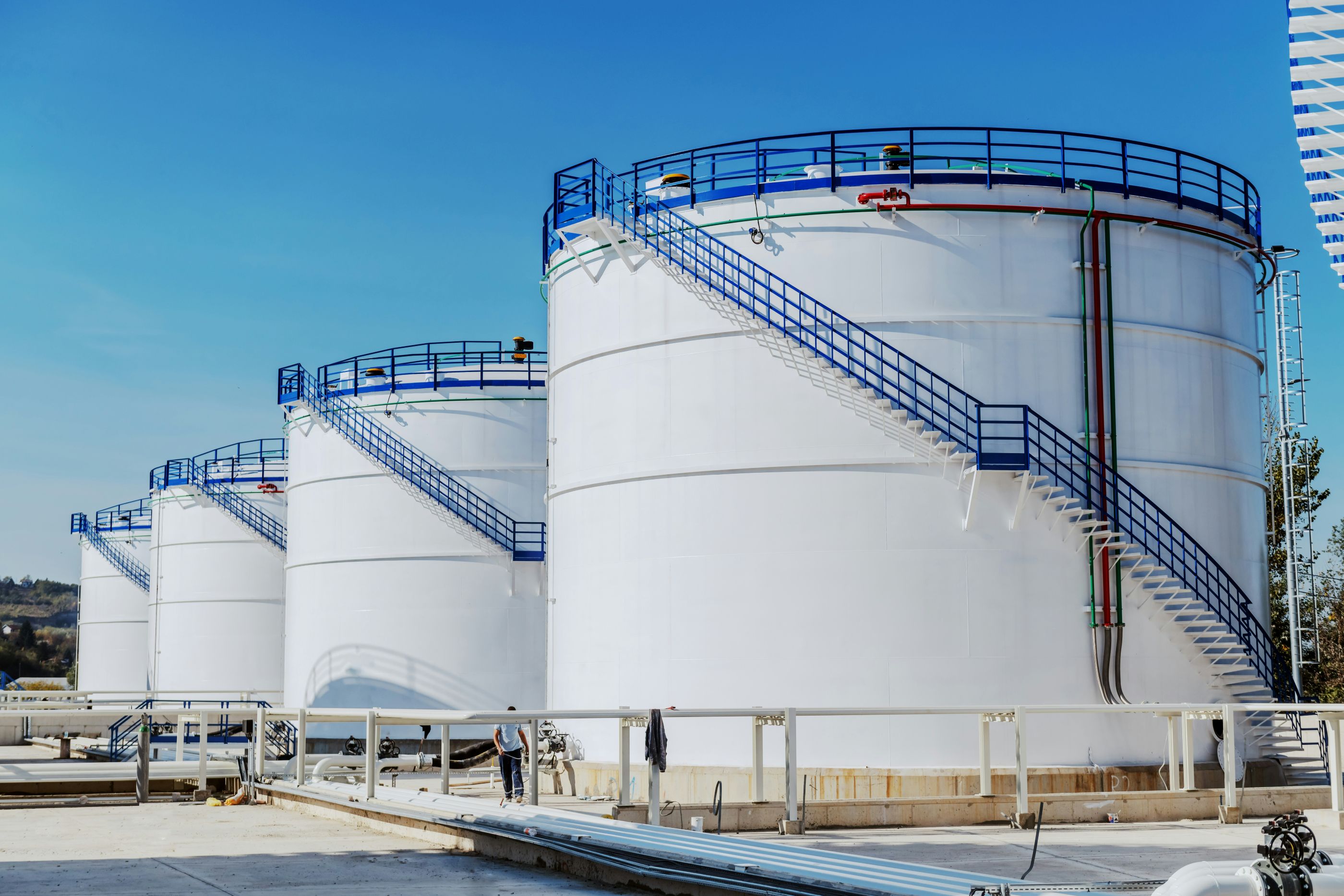 Row of large white cylindrical storage tanks with blue railings and staircases at an industrial facility under a clear blue sky.

