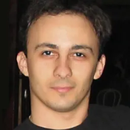 Portret apropiat al unui bărbat tânăr cu păr scurt și întunecat, cu un zâmbet subtil, pe fundal întunecat.