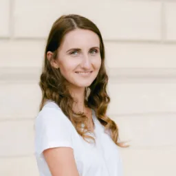 Femeie tânără cu păr șaten ondulat și bluza albă zâmbind ușor, pe fundal deschis.