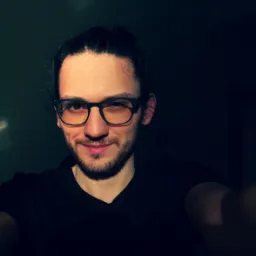 Bărbat tânăr cu păr lung și ochelari, zâmbind într-un mediu întunecat.