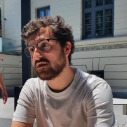 Bărbat cu păr creț și barbă, purtând ochelari și un tricou alb, stând în aer liber lângă o clădire.