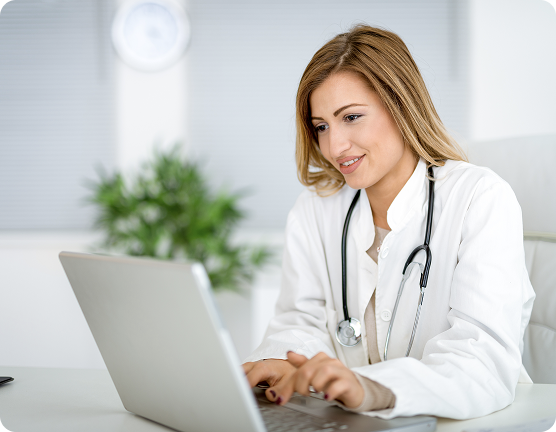 Medic specialistă zâmbitoare, în halat alb și cu stetoscop, lucrând pe un laptop într-un cabinet modern.