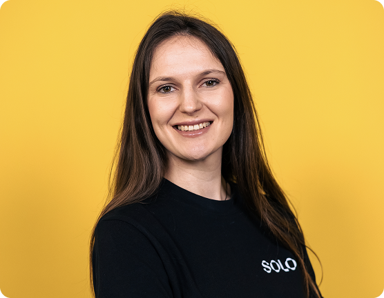 Portret al unei femei zâmbitoare cu păr lung și brunet, purtând un tricou negru cu textul 'SOLO' pe fundal galben.