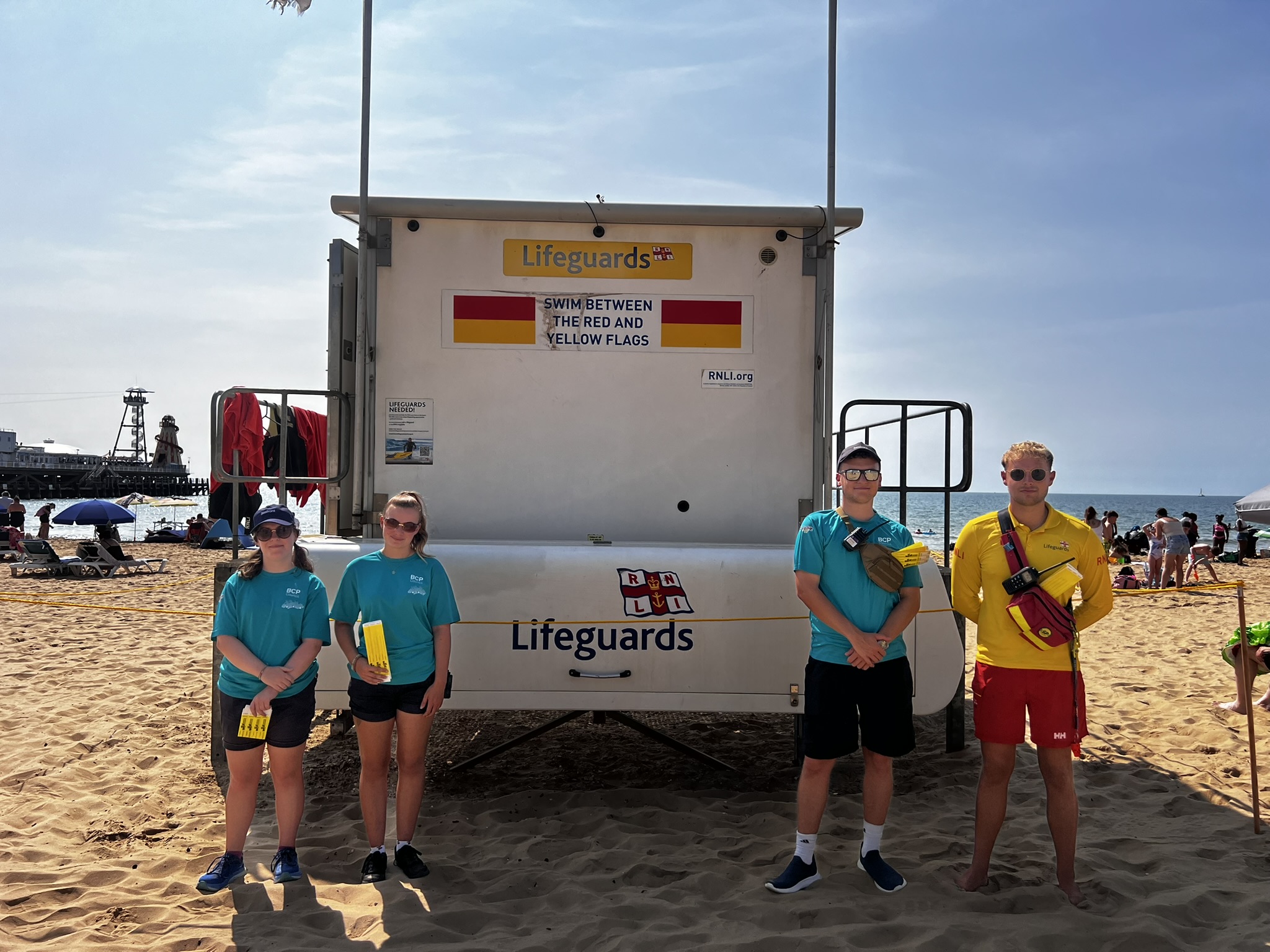 BCP Council Seafront Team and RNLI Lifeguard standing in front of Lifeguard Tower at Bournemouth Beach West with Yellow LV= KidZone Wristbands.