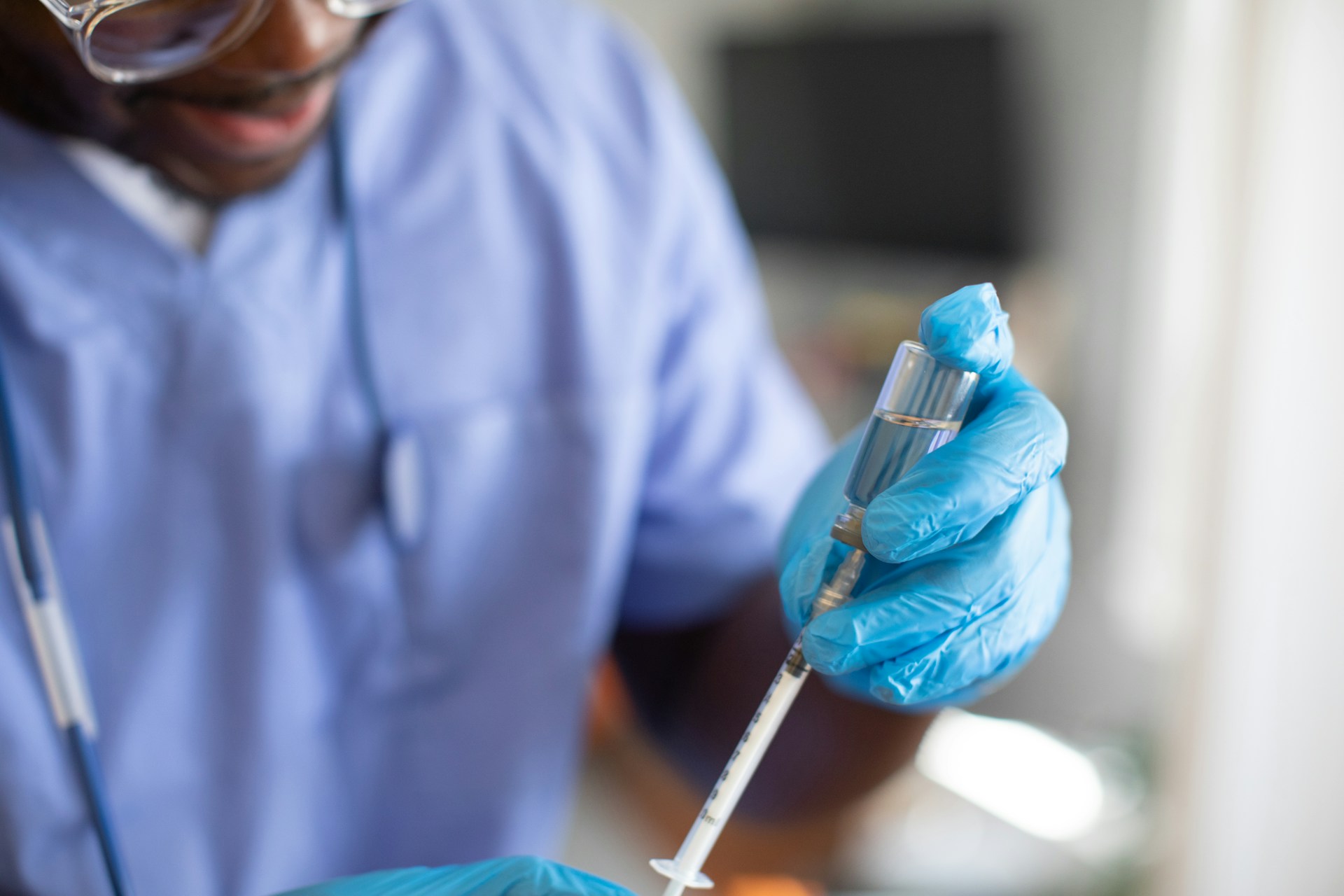 Healthcare professional in blue scrubs and gloves preparing a syringe with a vaccine.