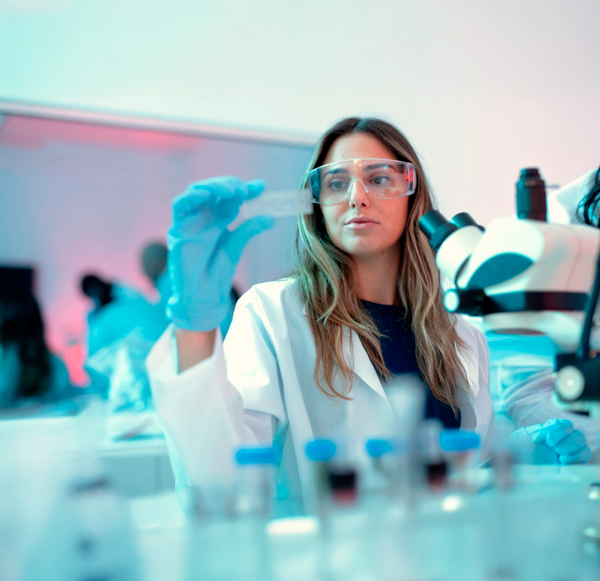 Female scientist in lab coat and safety glasses examining a microscope slide in a laboratory.