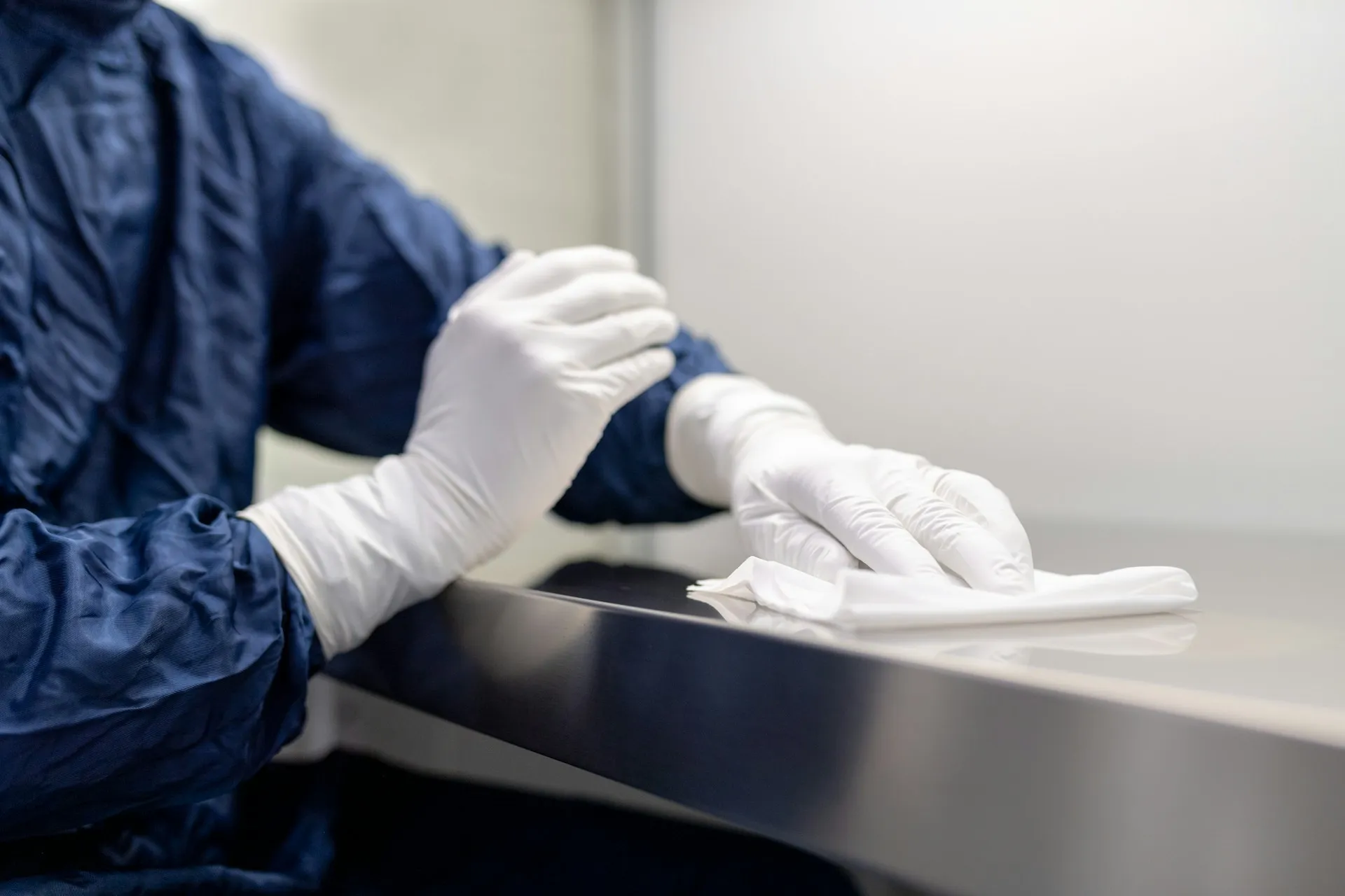 Person wearing white gloves and a blue protective suit cleaning a stainless steel surface with a white cloth.