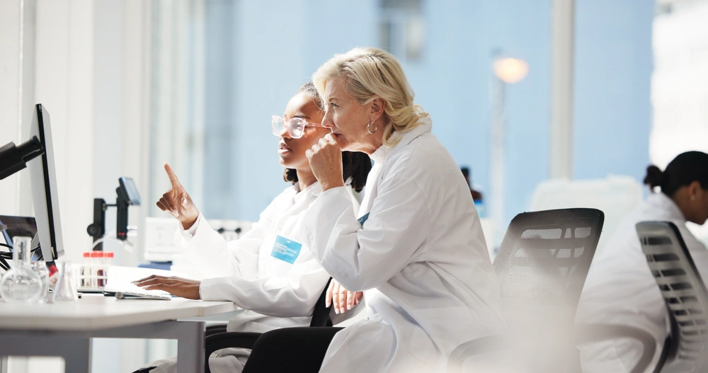 Two female scientists in white lab coats looking at a computer screen in a modern laboratory.
