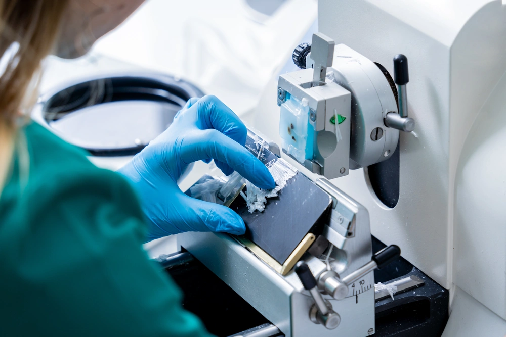 Male scientist in white lab coat looking through a microscope in a laboratory with various bottles and test tubes.