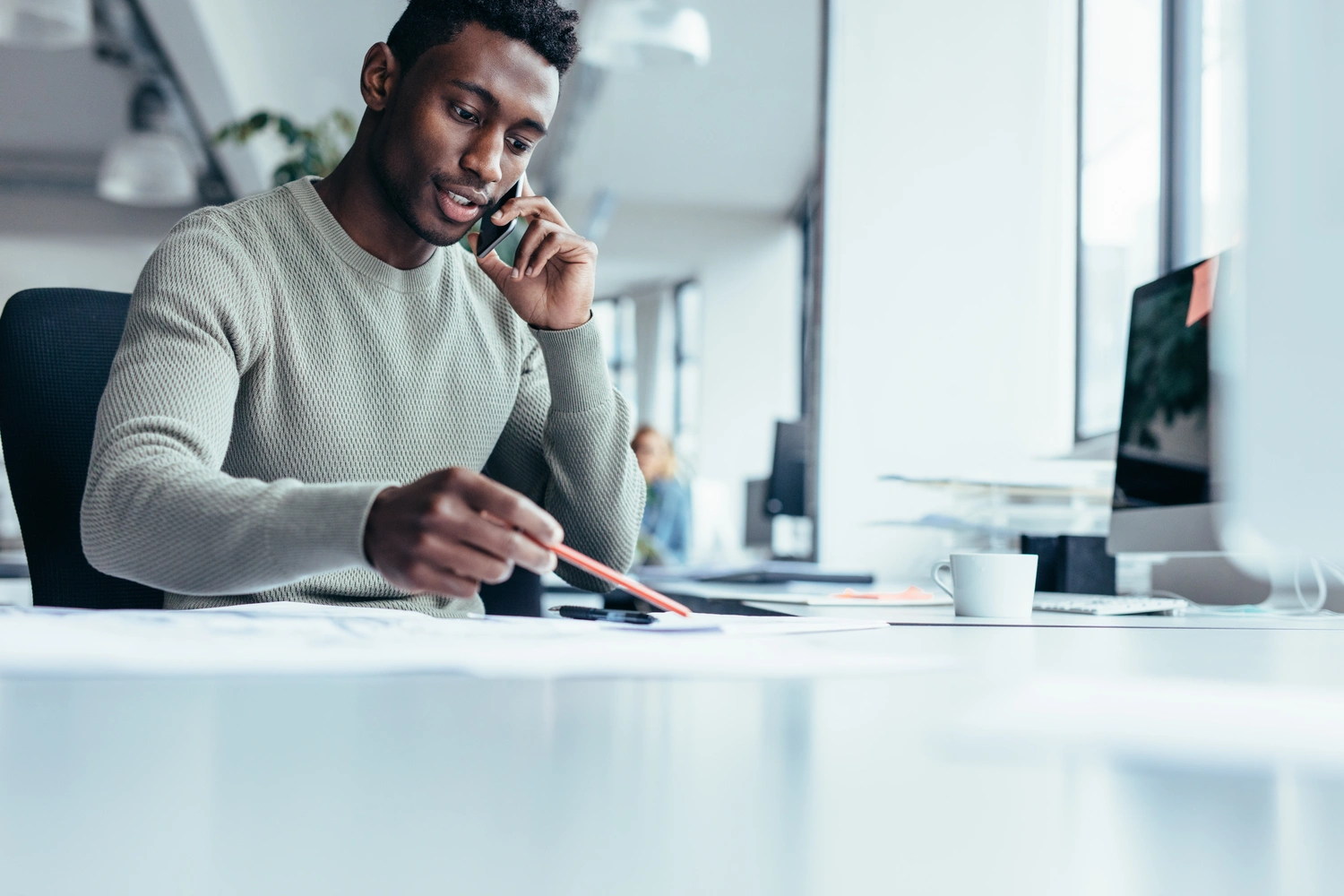 Man talking on phone while pointing at papers with a pencil at a desk in a bright office.