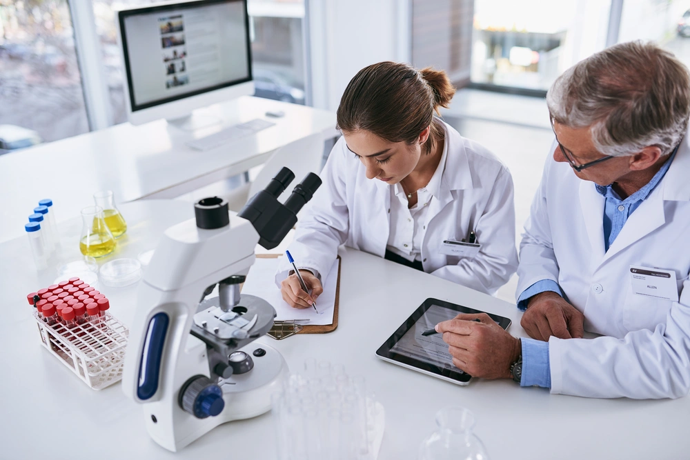 Male scientist in white lab coat looking through a microscope in a laboratory with various bottles and test tubes.