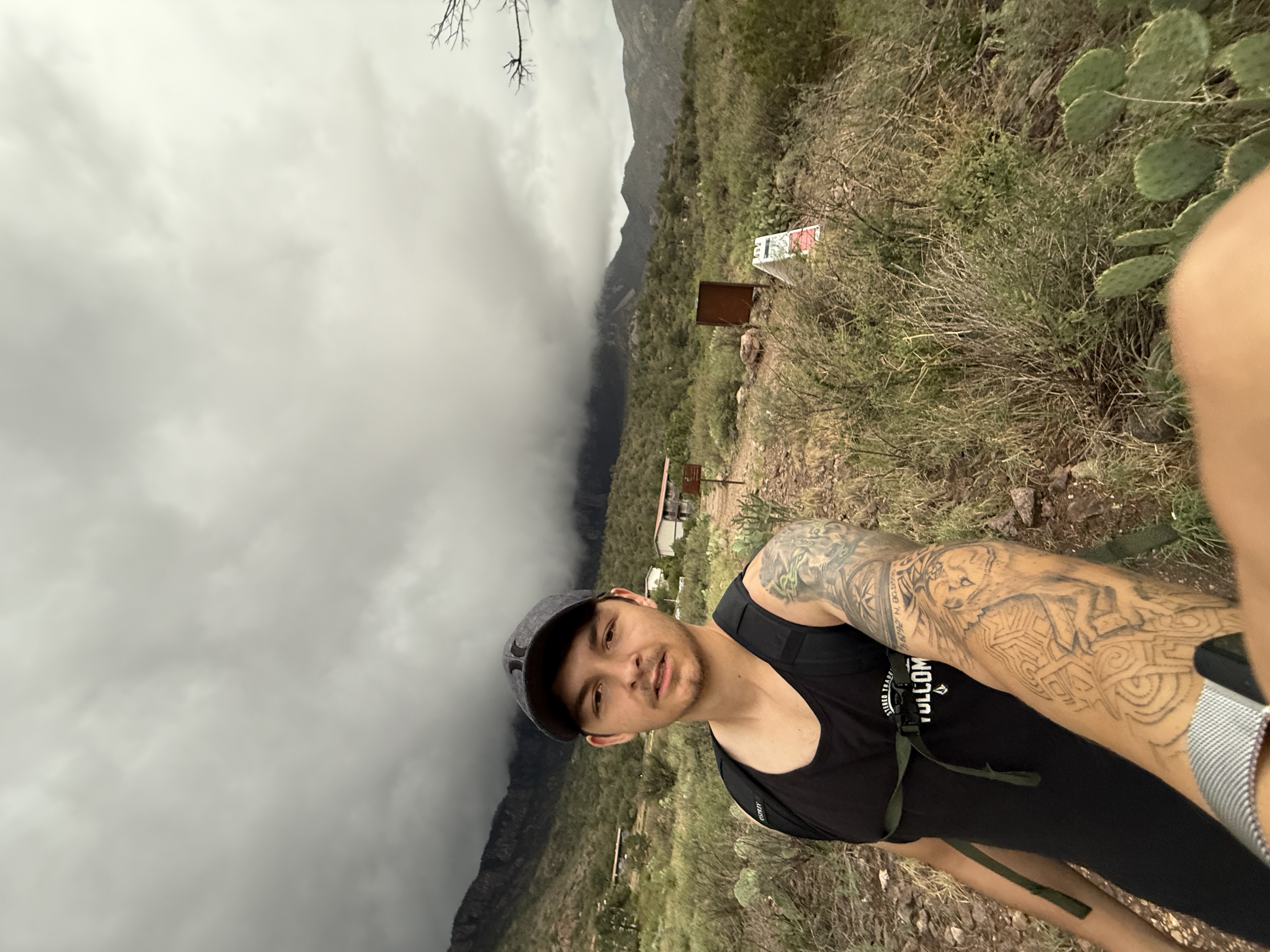 Man with a tattooed arm wearing a black tank top and cap taking a selfie on a rocky path with green shrubs, cacti, and cloudy mountains in the background.