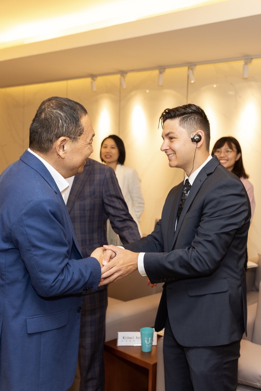 Two men in suits smiling and shaking hands indoors with two women standing in the background.