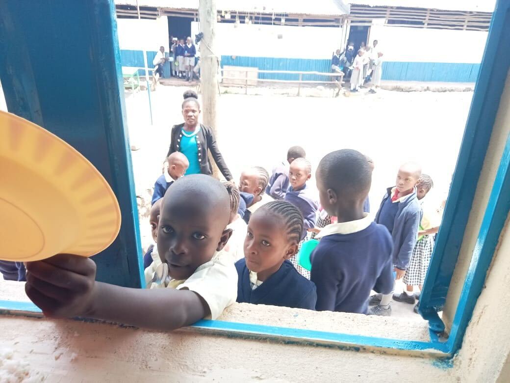 Children in school uniforms standing outside a window, one boy holding a yellow plate toward the camera.