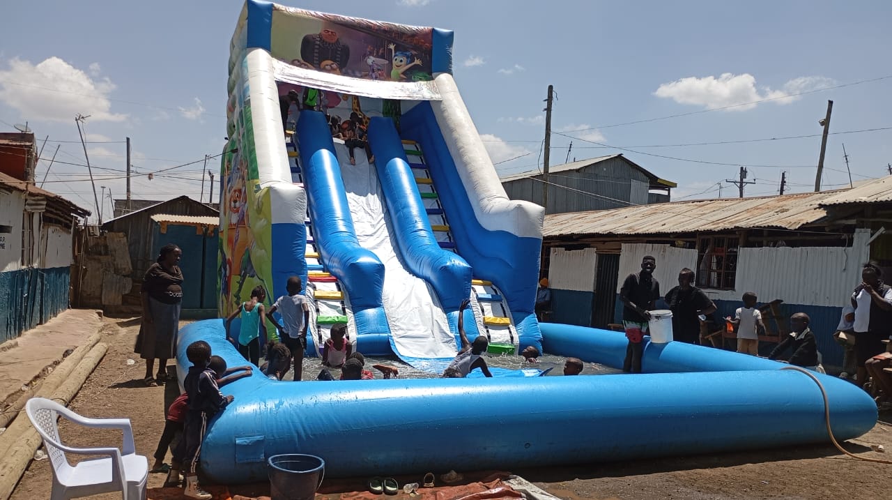 Children playing on a large blue inflatable water slide with a splash pool at the bottom, set outdoors in a dusty neighborhood.
