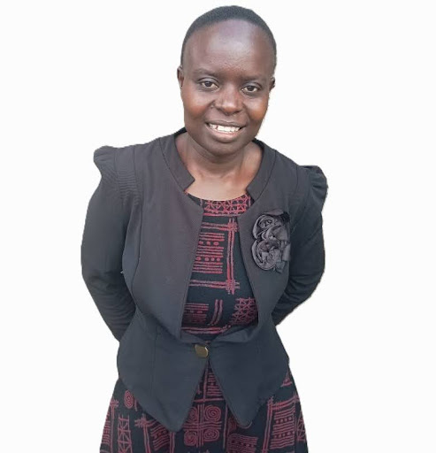 Smiling woman wearing a black blazer over a patterned dress with hands behind her back against white background.