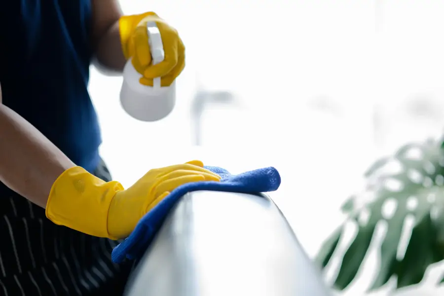 Person wearing yellow gloves sprays cleaner and wipes a surface with a blue cloth; plant blurred in background.
