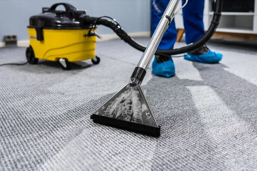 Carpet cleaner with yellow tank and hose being used to deep clean grey carpet; person wearing blue shoe covers.