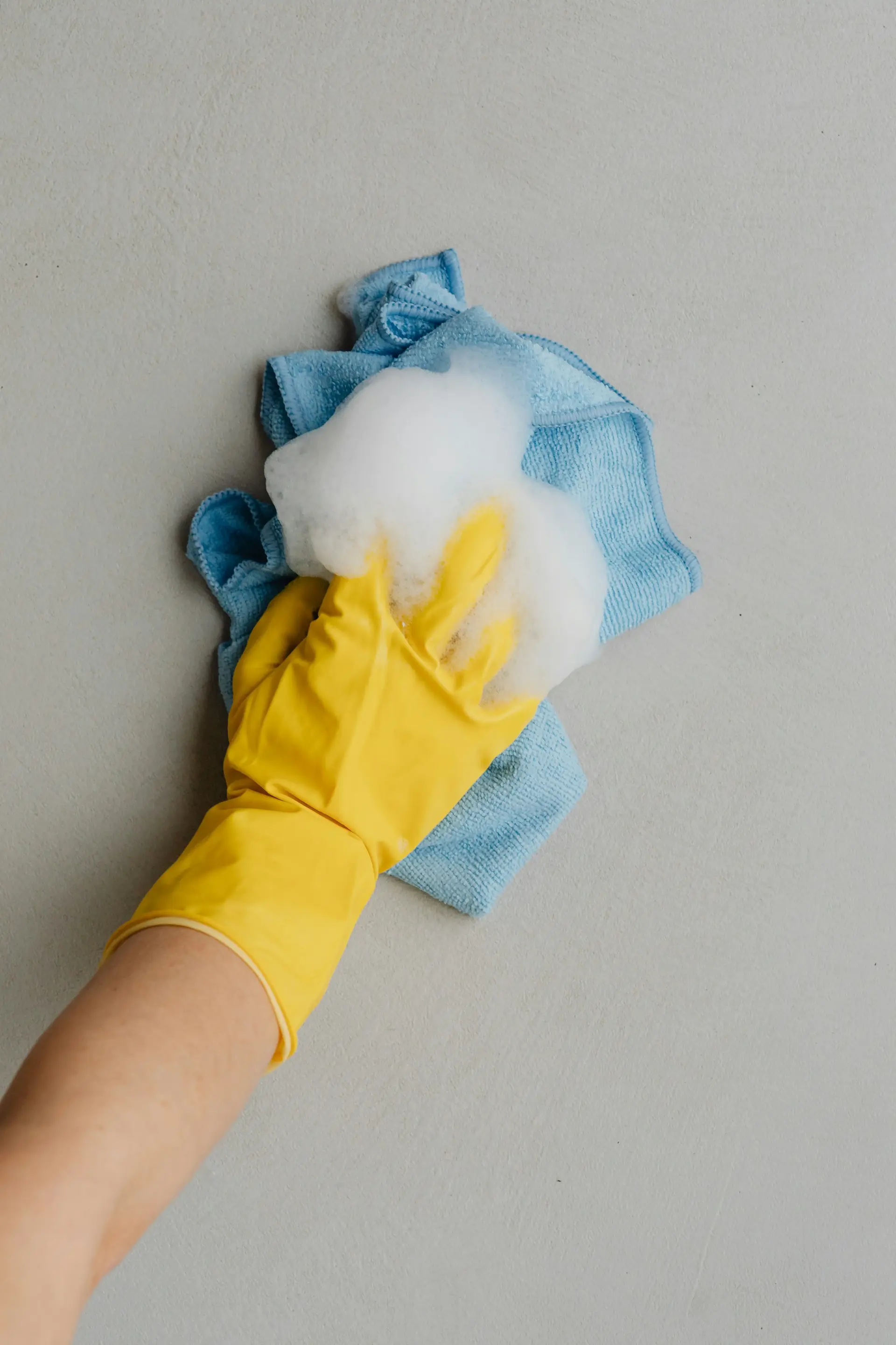 Yellow-gloved hand cleaning a grey surface with a blue cloth and soap suds.