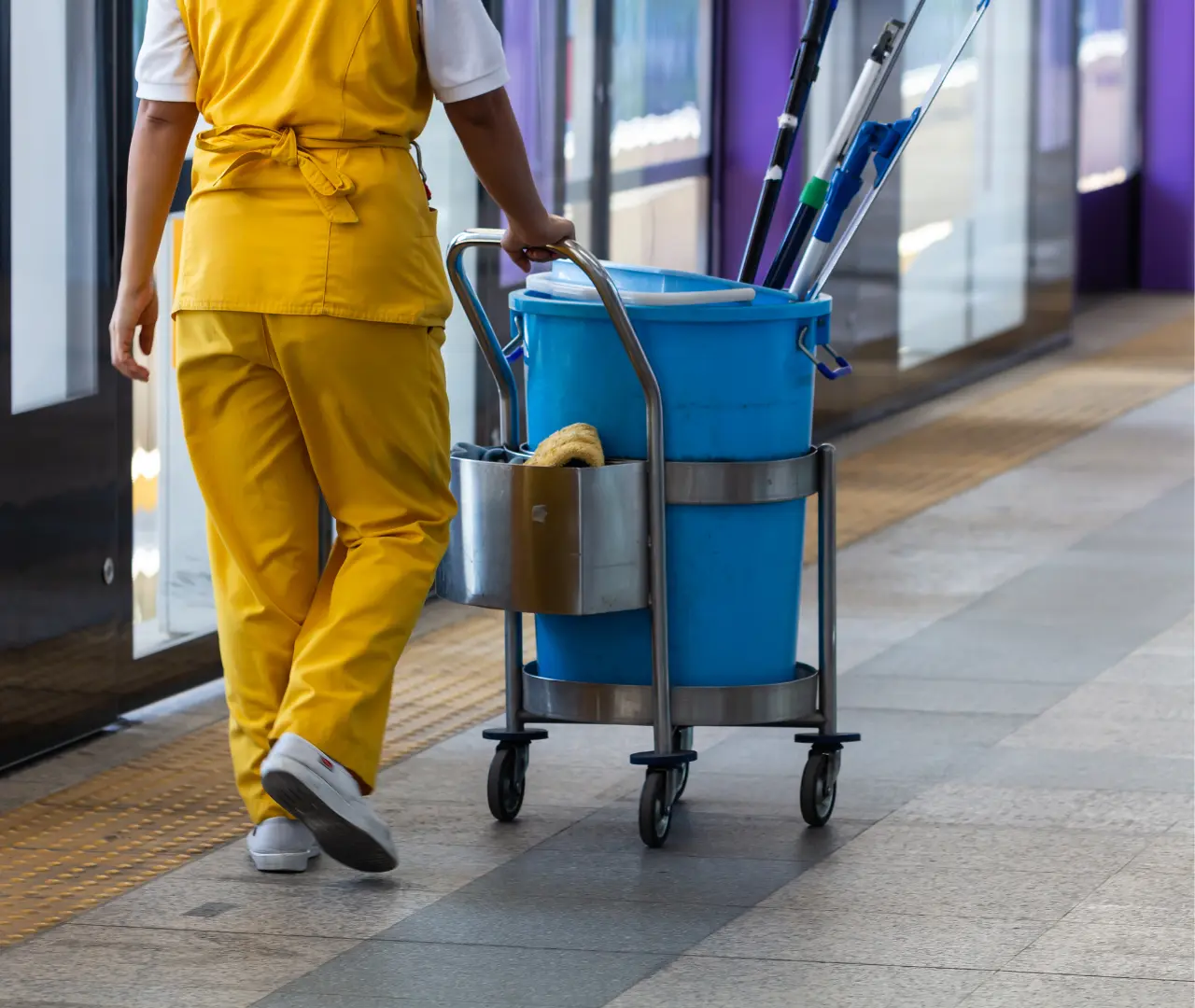 Cleaner in yellow uniform pushes a blue cleaning cart with mops and supplies along a tiled hallway.