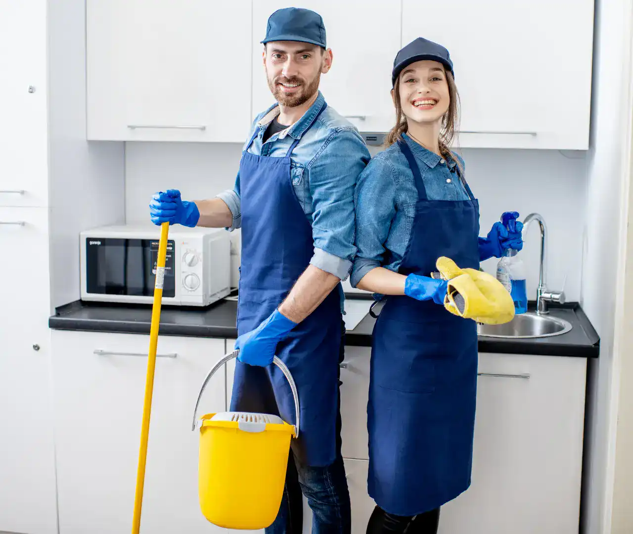 Two smiling cleaners in blue aprons and gloves stand in a kitchen, holding a mop, yellow bucket, and cleaning cloth.