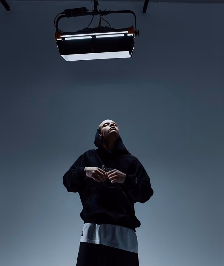 Person wearing a black hoodie looking up toward a large overhead studio light in a dark room.