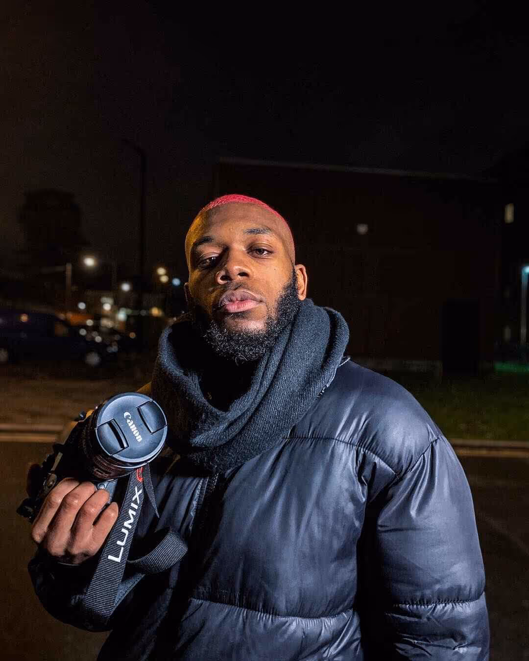 Man with dyed pink hair and beard wearing a black puffer jacket and scarf holding a camera at night outdoors.