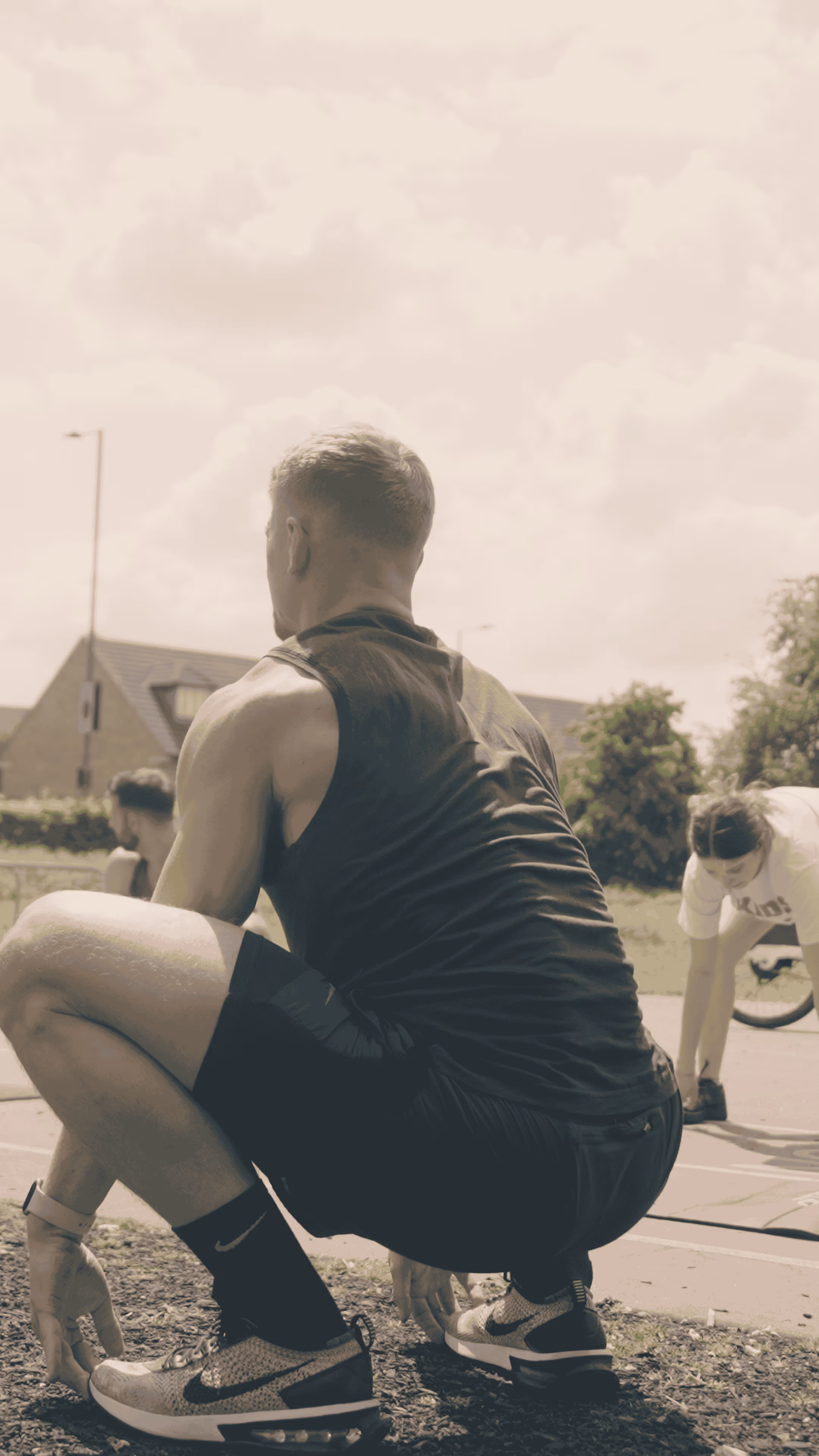 Person in black athletic wear squatting outdoors with another person stretching in the background near grassy area and houses.
