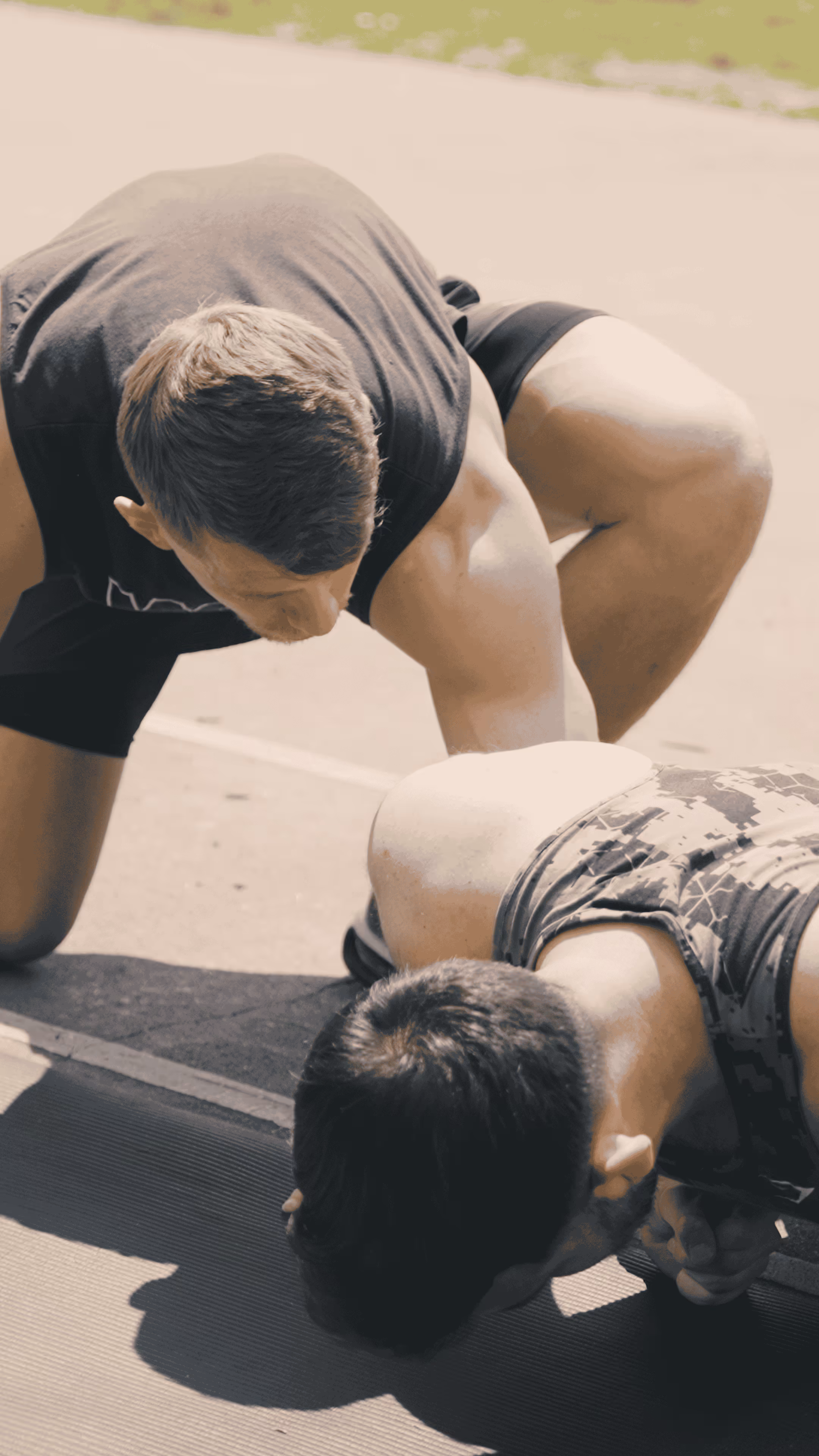 A man in a black sleeveless shirt coaches another man doing a push-up on a mat outdoors.