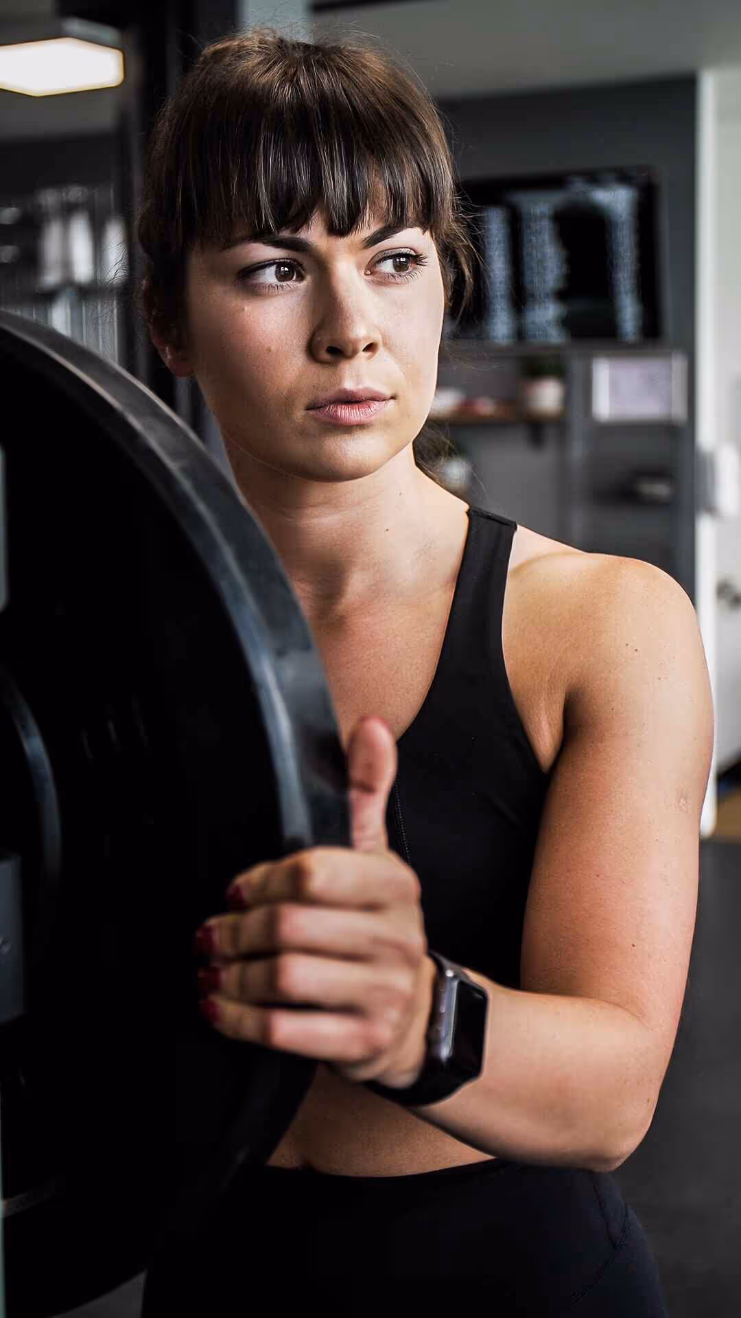 Woman in black sportswear holding a weight plate in a gym, focused and looking to the side.