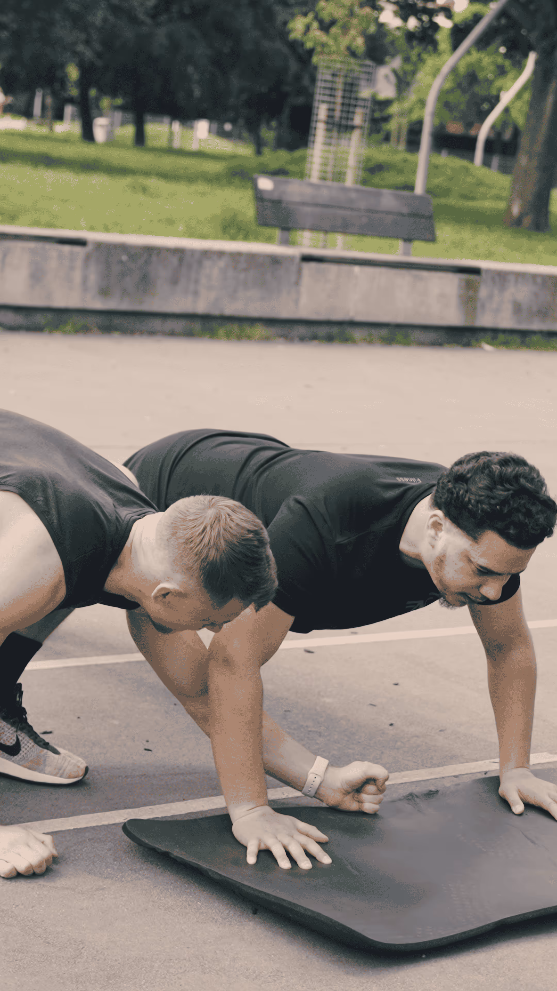 Two men doing push-ups on a black exercise mat outdoors near a park bench.