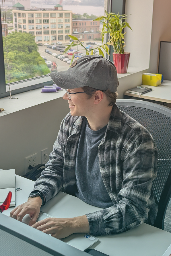 Person wearing glasses, a gray cap, and a plaid shirt sitting at a desk and working on a laptop near a window with a view of buildings and parked cars.