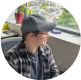 Young man wearing glasses, a dark cap, and a plaid shirt looking out of a window.