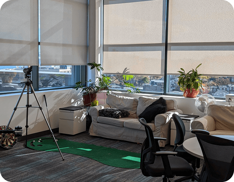 Sunlit GreenSight Boston office lounge with beige sofas, potted plants on windowsill, a tripod standing on a putting green carpet, and black office chairs around a table.