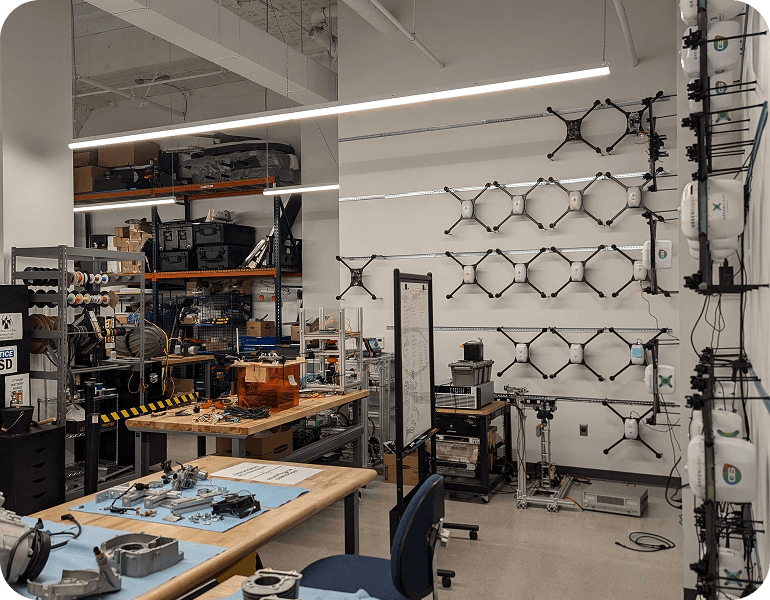 Workshop room in GreenSight Boston Office with multiple assembly tables, drone frames mounted on the wall, and various tools and equipment organized on shelves and tables.