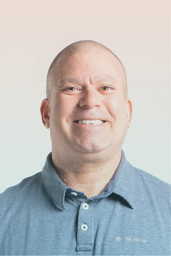 Smiling bald man wearing a blue Columbia polo shirt against a gradient beige to light blue background.