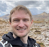 Man smiling outdoors in a rocky mountainous landscape under a cloudy sky.