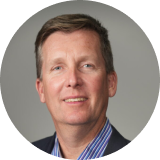 Portrait of a middle-aged man with short brown hair wearing a blue striped shirt and dark blazer against a gray background.