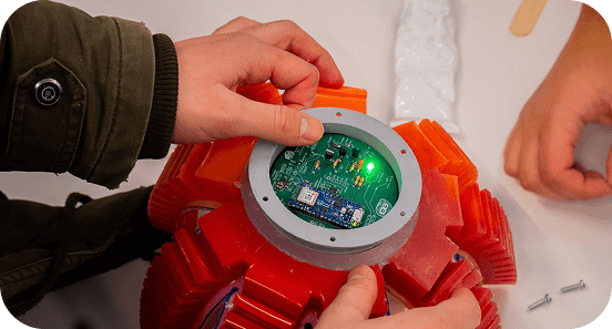 Hands assembling a red mechanical device with a visible circuit board and a glowing green LED light.