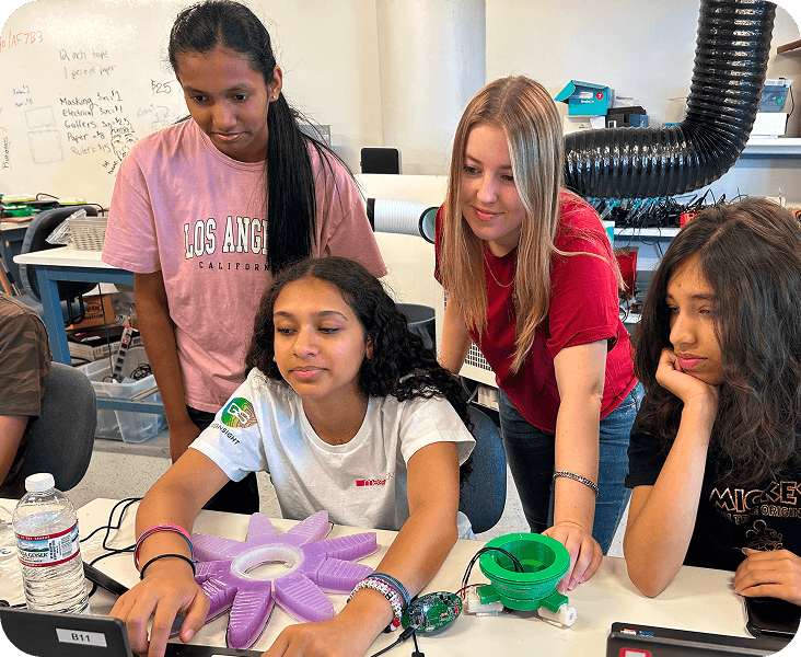 Four teenage girls collaborate around a table with electronic devices and a purple star-shaped object, engaged in a STEM activity in a classroom.