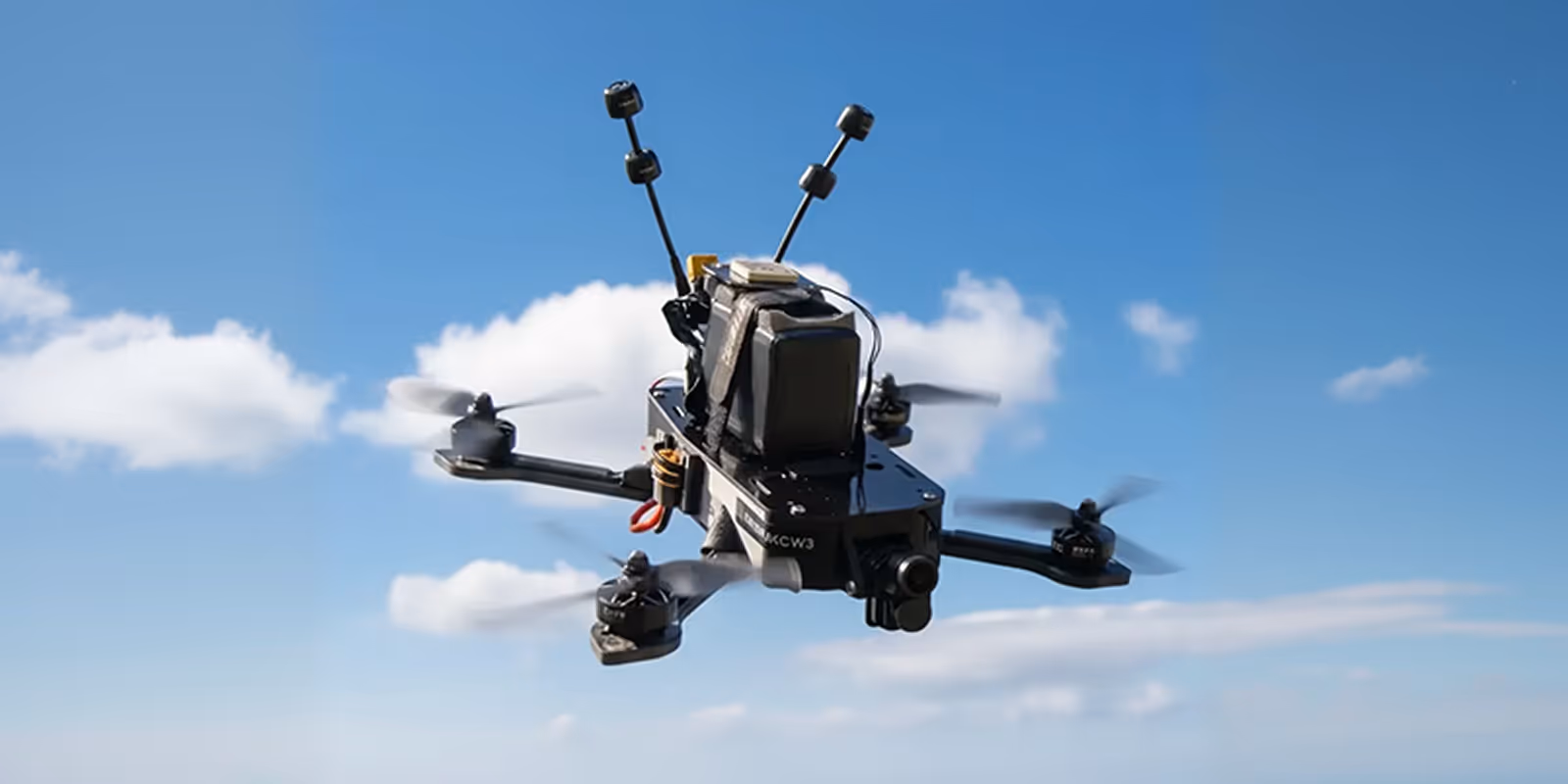 Black quadcopter drone flying against a blue sky with white clouds.