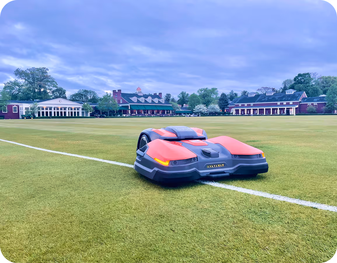 Husqvarna robotic lawn mower operating on a large, well-maintained sports field with buildings and trees in the background under a cloudy sky.