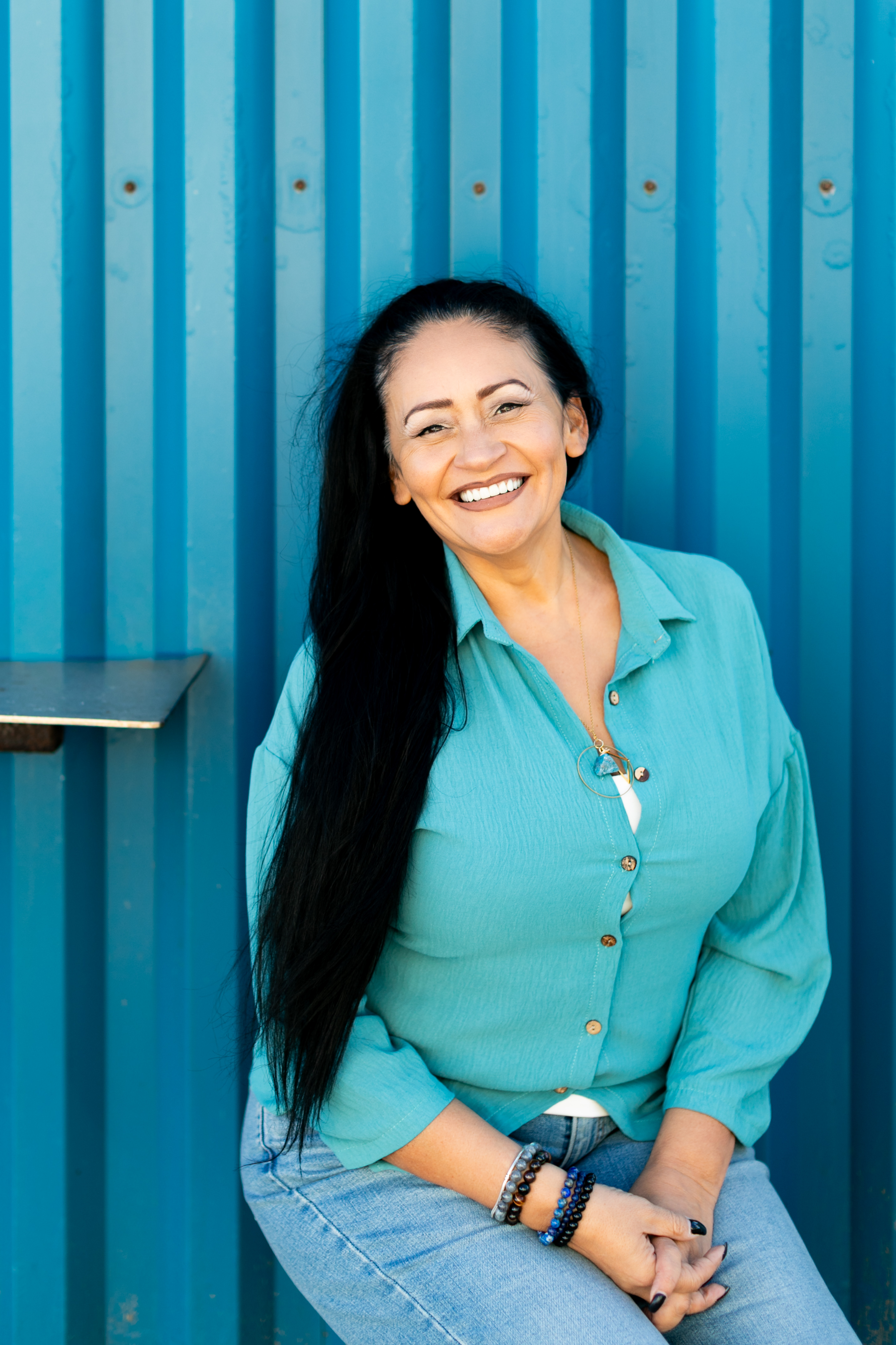 Smiling woman with curly blonde hair sitting on a couch holding sunglasses and a book.