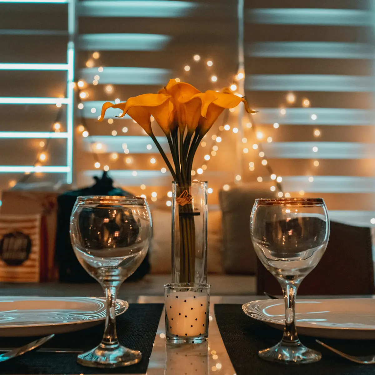 Romantic dinner setup with wine glasses, yellow flowers, candle, and fairy lights.