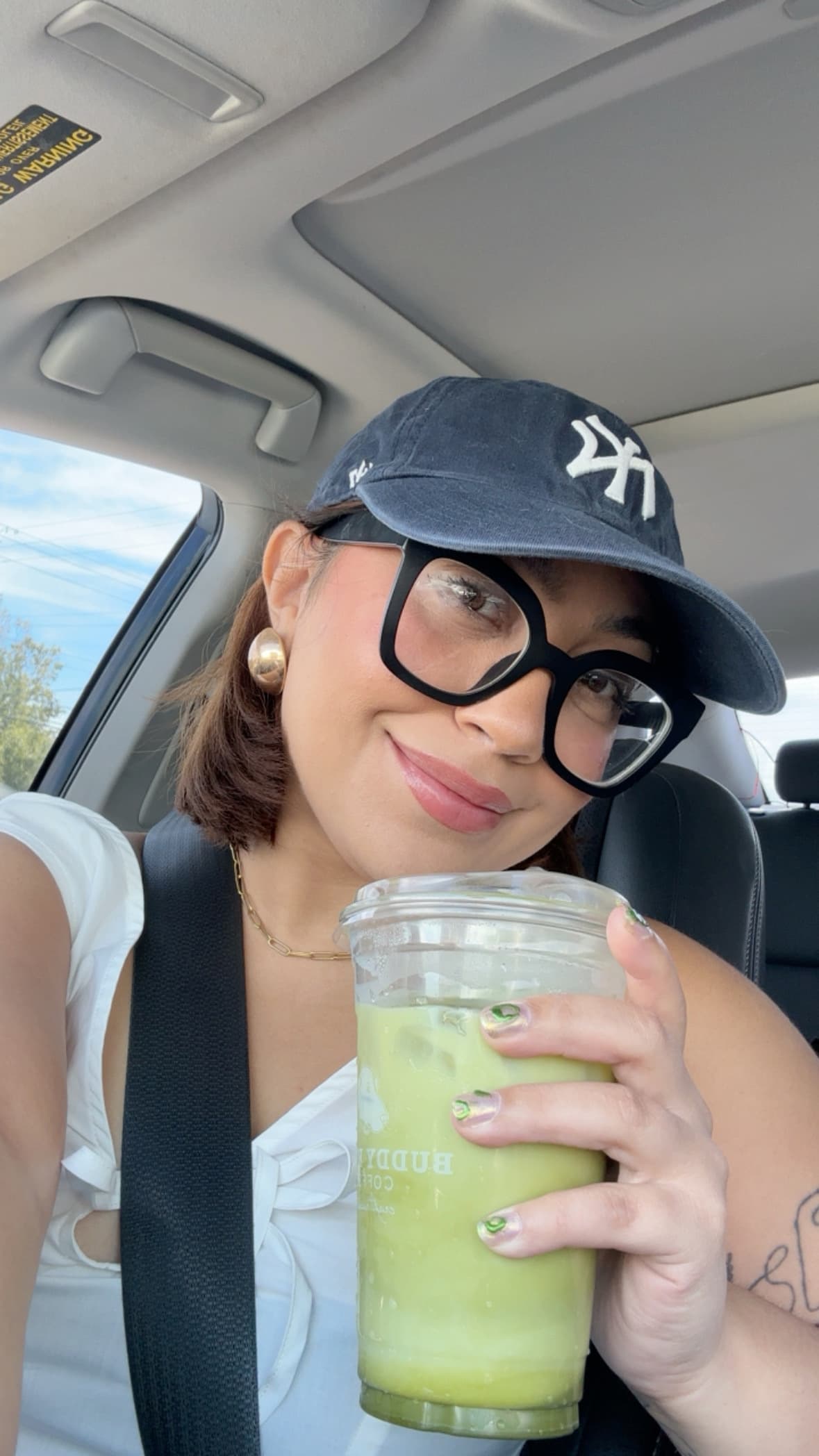 Girl wearing a hat and glasses taking a selfie with an iced matcha latte