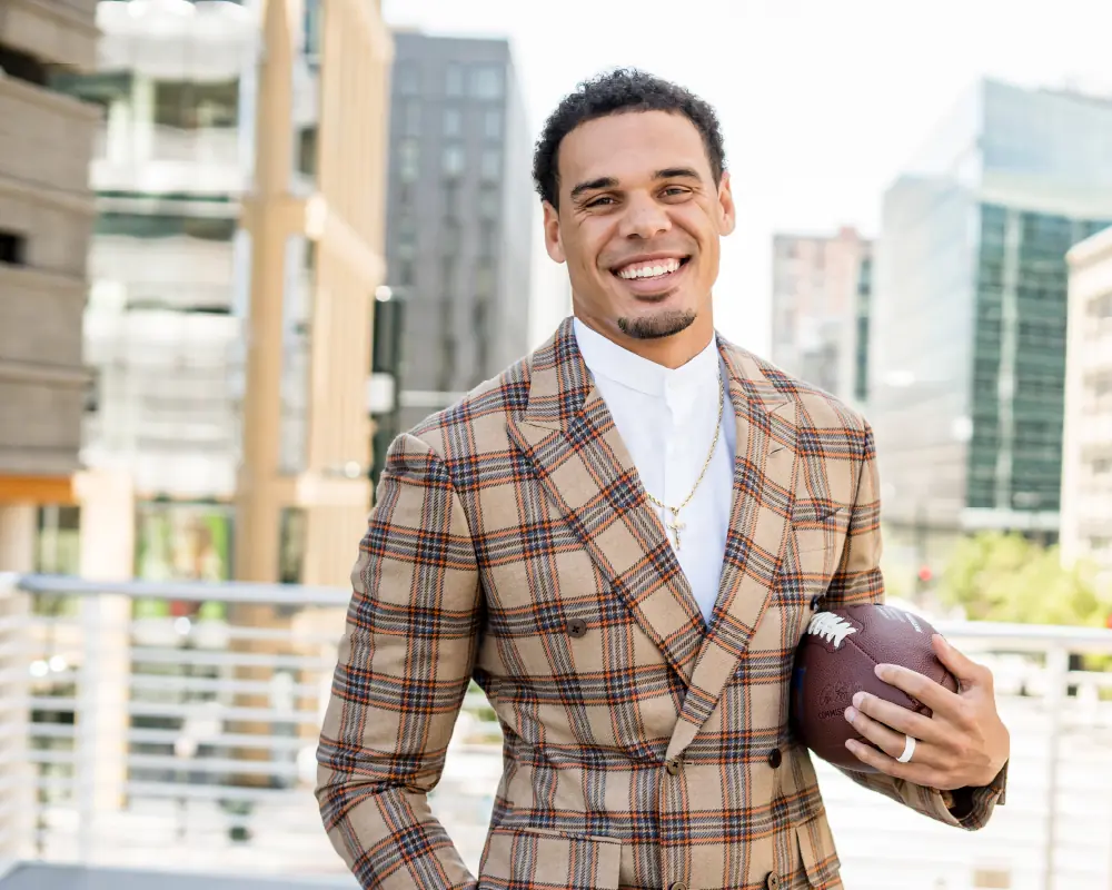 Professional football player Justin Simmons smiles while standing outdoors in a plaid suit, holding a football with city buildings in the background.