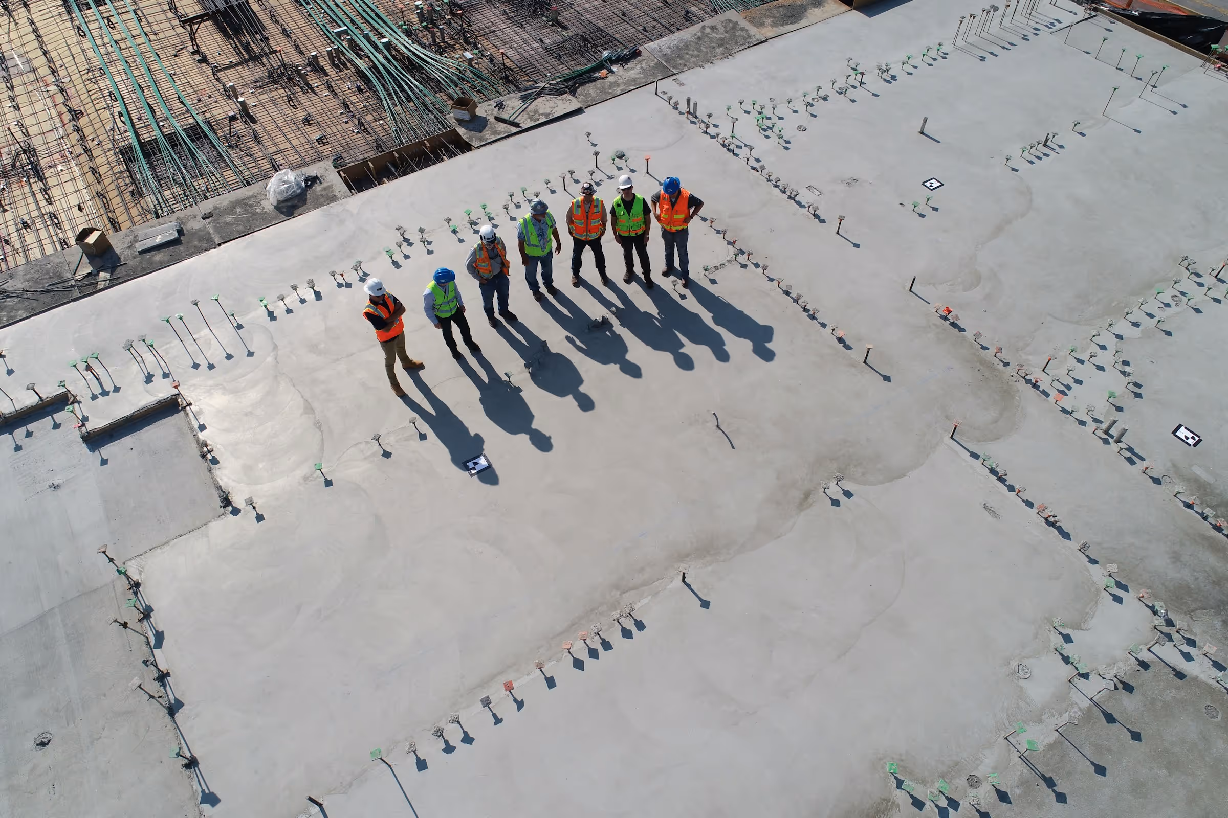 Eine Gruppe von zehn Bauarbeitern in Schutzkleidung steht auf einer frisch gegossenen Betonfläche. Ihre Schatten fallen auf den glatten Boden, während im Hintergrund Baustellenmaterialien zu sehen sind.